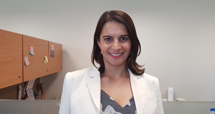 A woman in a white blazer smiles while standing in an office setting.